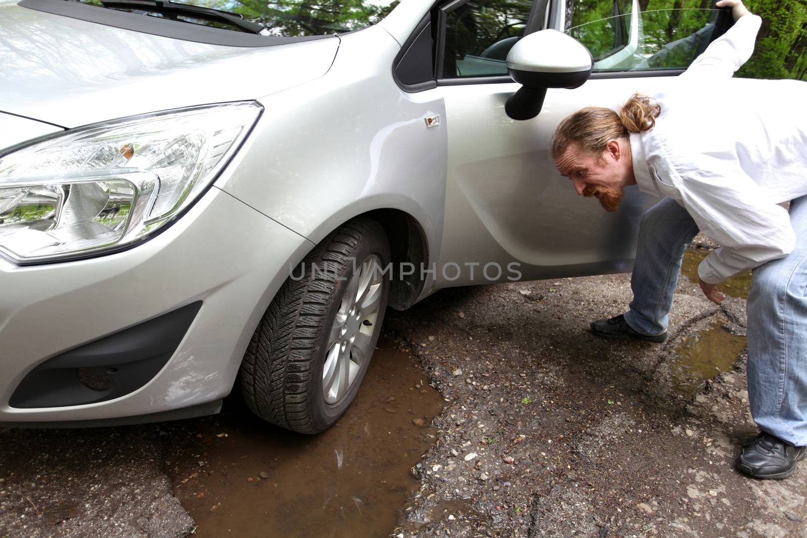 Upset driver man in front of automobile watching damaged car of road full of cracked potholes in pavement