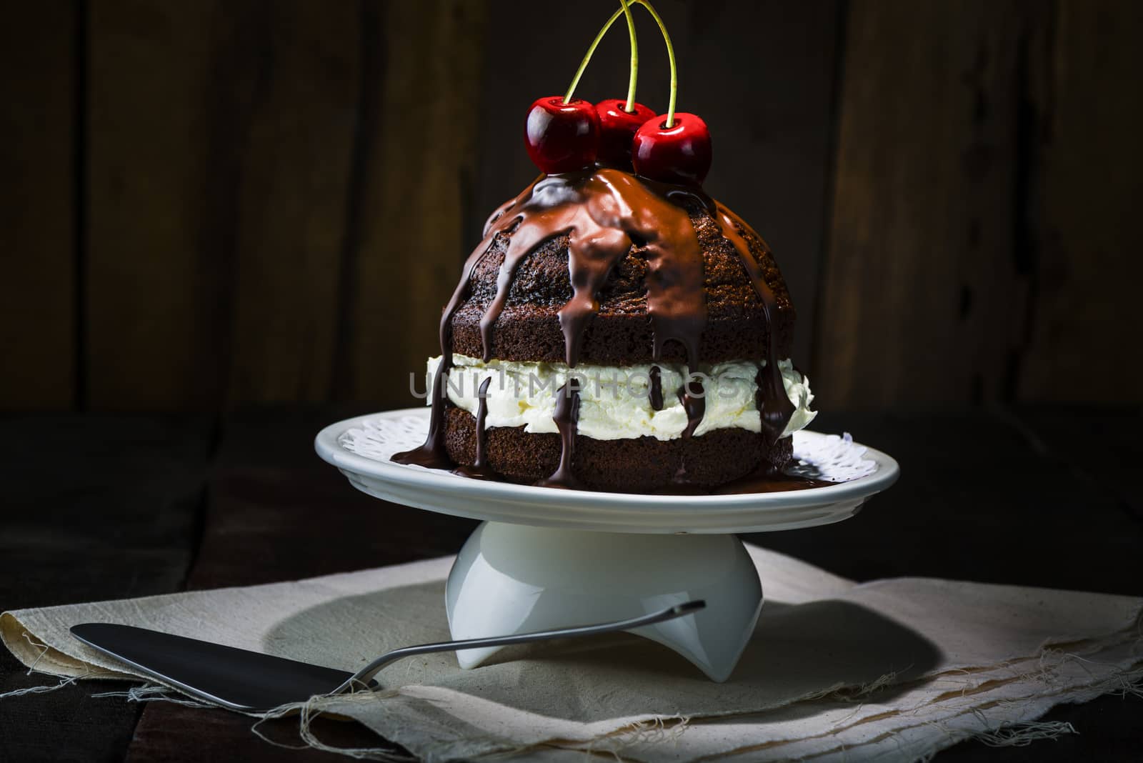 Delicious Chocolate Cake, Served on White Porcelain Tray, with Red Cherries on Top. Place on Top of Napkin at the Wooden Table with Spoon.