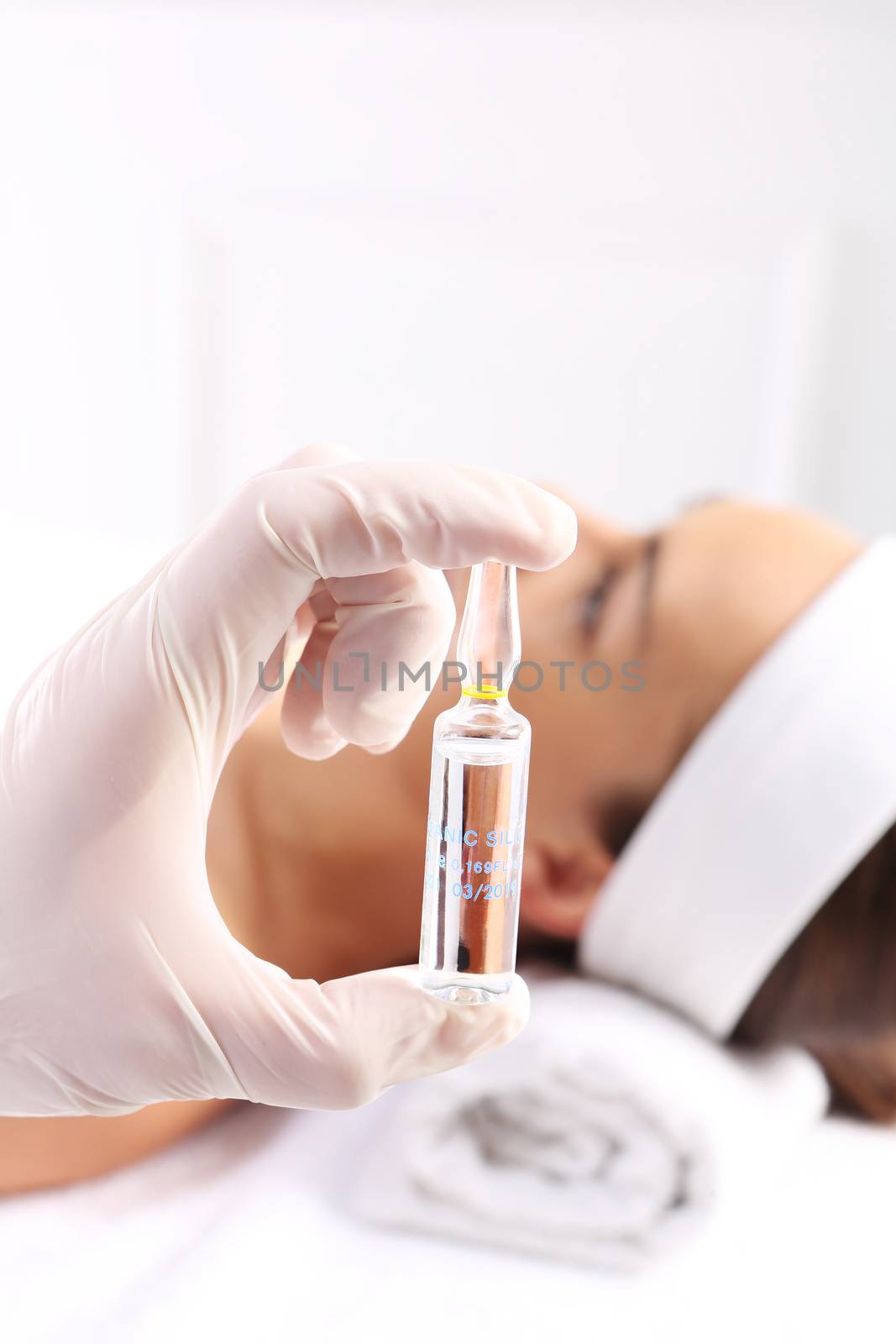 Gloved hand medical shows ampoule with a cosmetic preparation, in the background a woman waiting for surgery by robert_przybysz