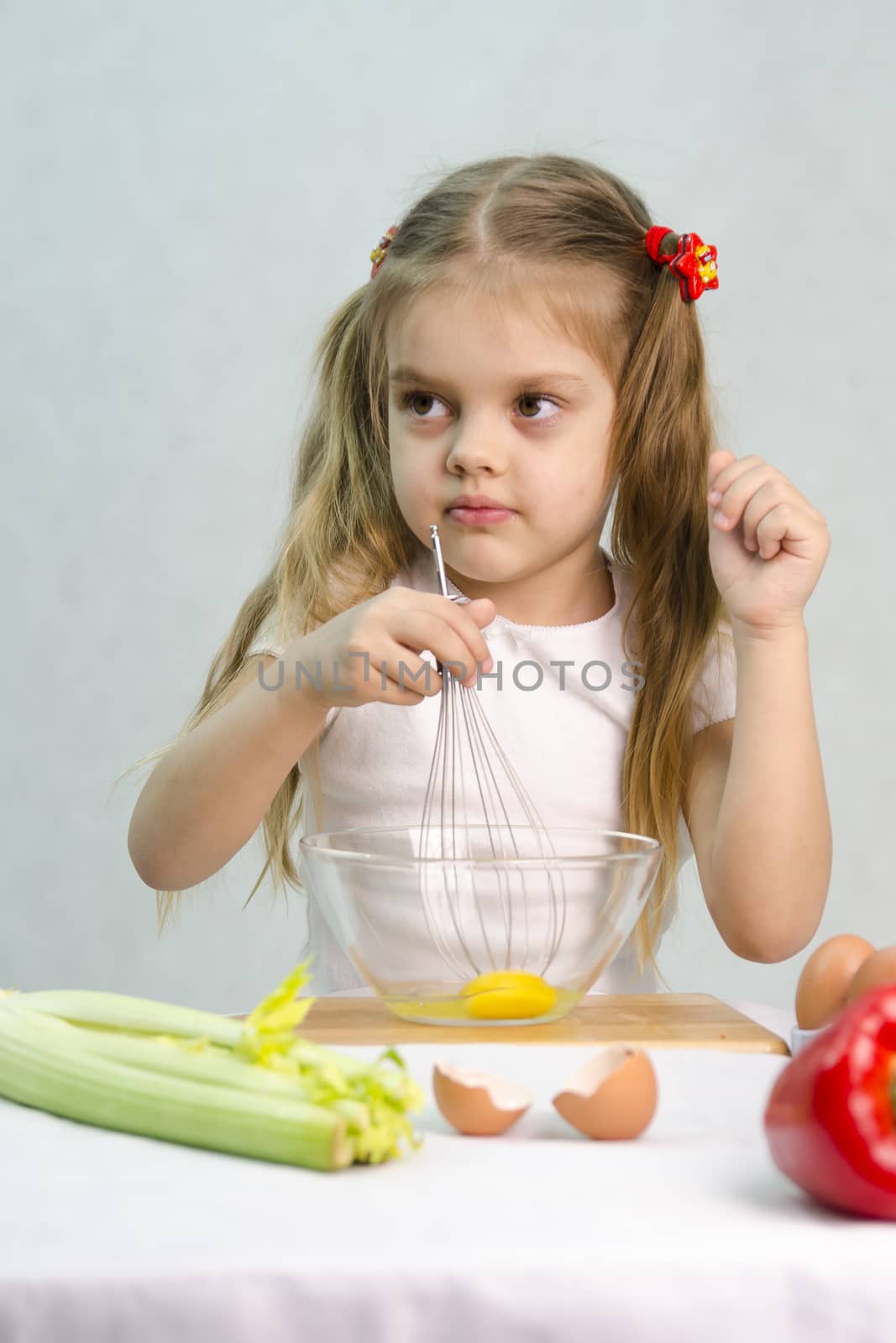Girl playing in a cook. Girl churn whisk the eggs in a glass bowl. At the forefront of the shell, celery, pepper. Studio. Light background.