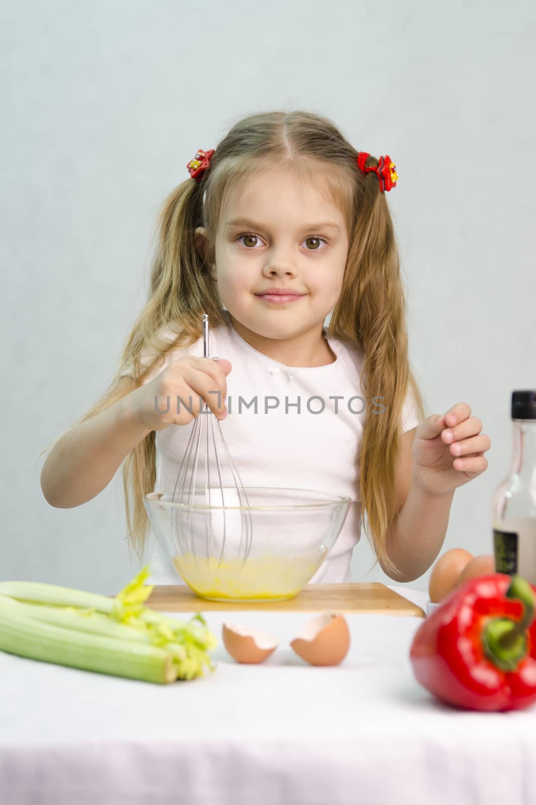Girl playing in a cook. Girl churn whisk the eggs in a glass bowl. At the forefront of the shell, celery, pepper. Studio. Light background.