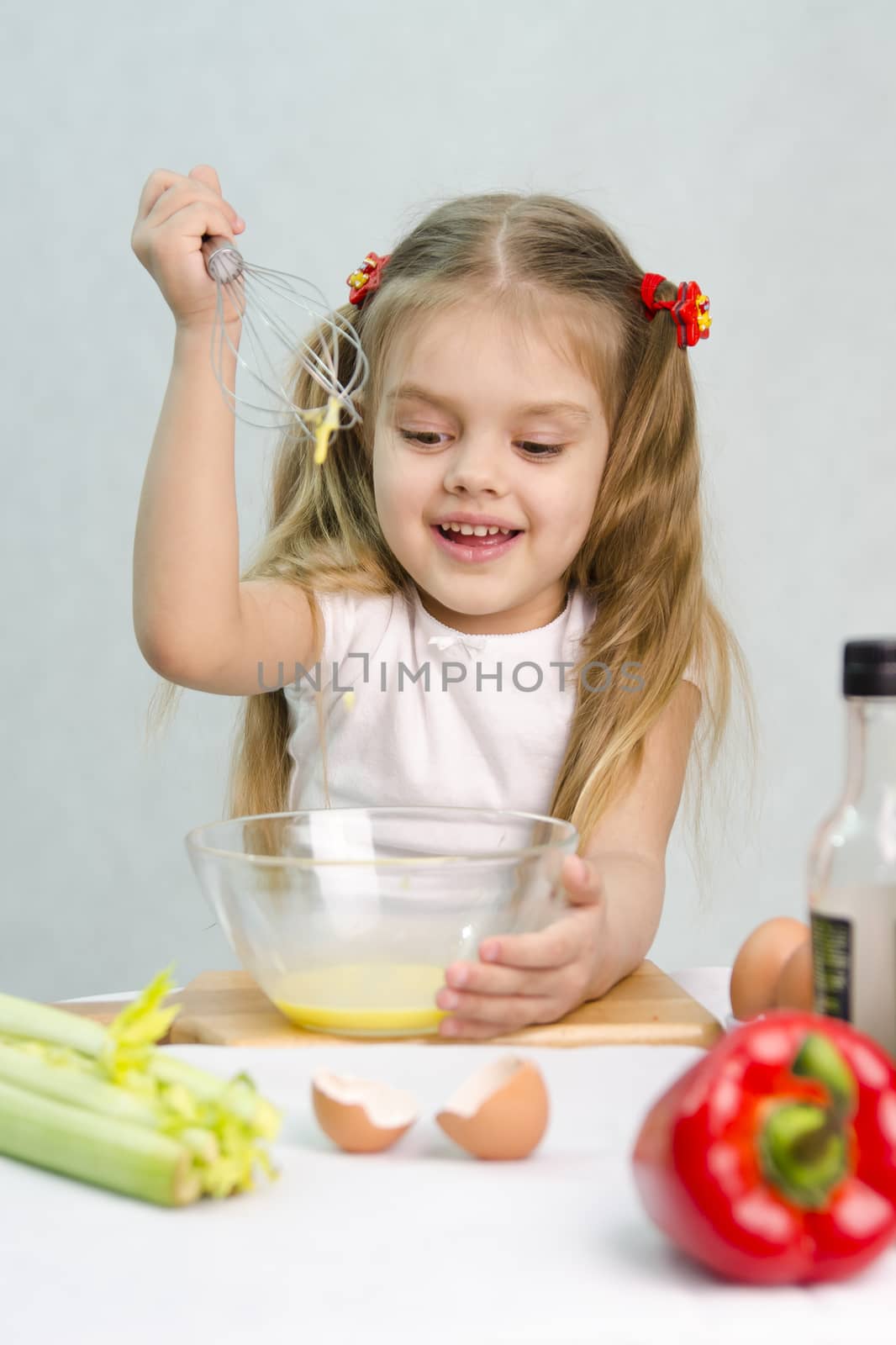 Girl playing in a cook. Girl churn whisk the eggs in a glass bowl. At the forefront of the shell, celery, pepper. Studio. Light background.