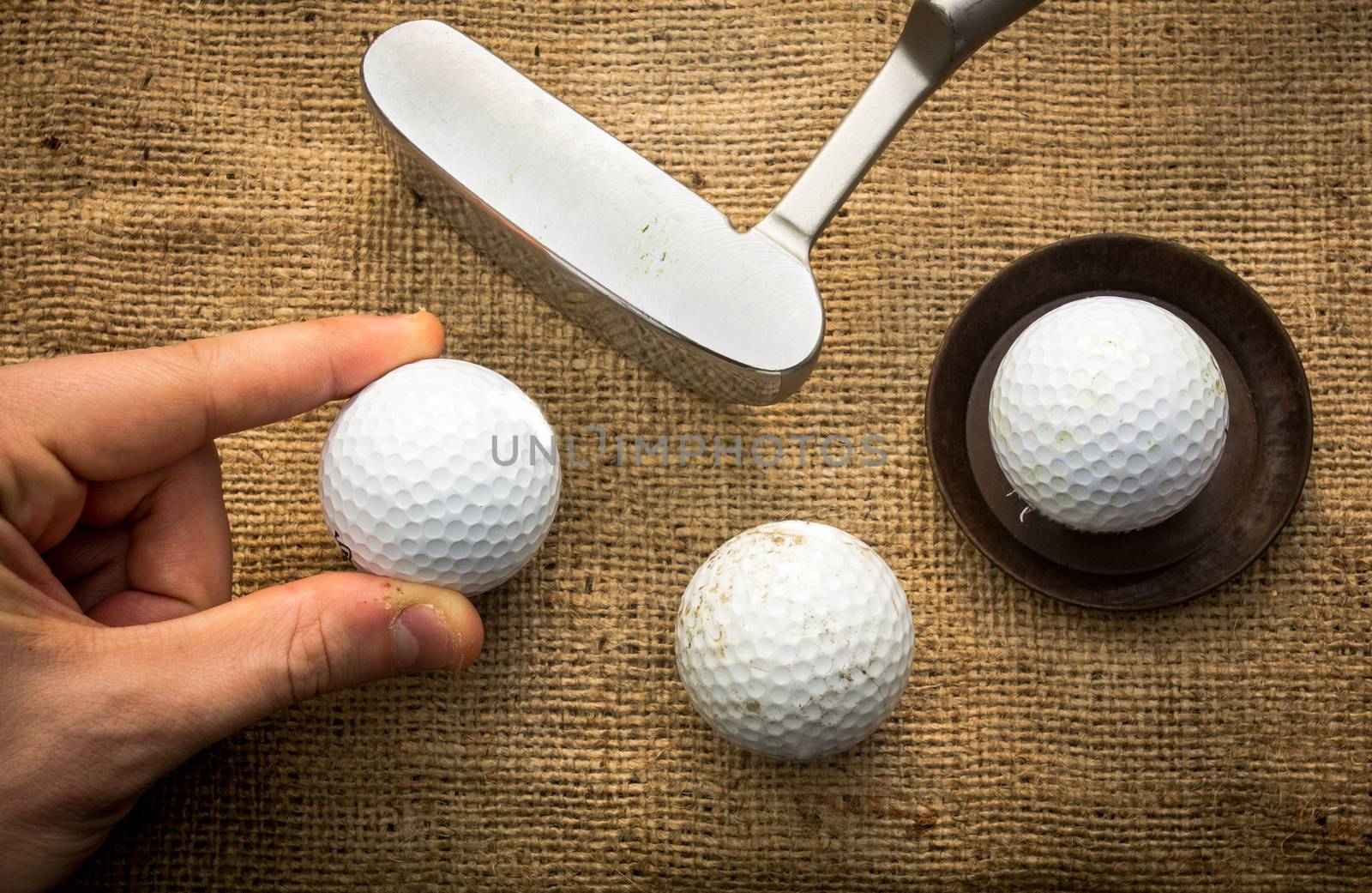 A hand holding a golf ball near a putter and two other golf balls