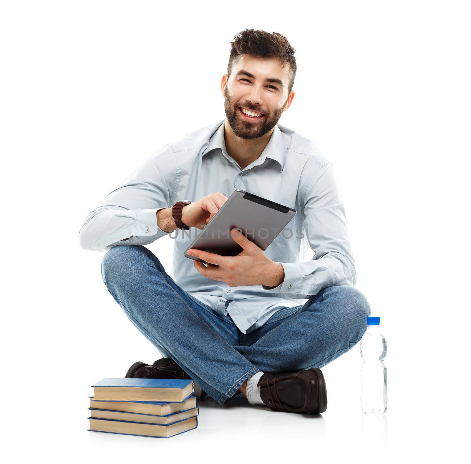 Young bearded smiling man holding a tablet with books and a bottle of water sitting on a white background