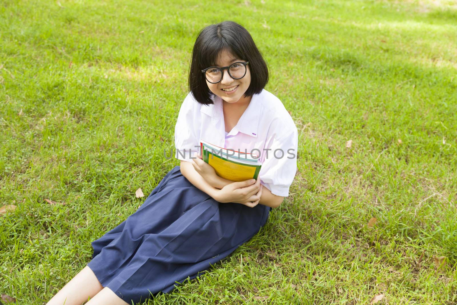 Schoolgirl holding books and smiling. Sitting on grass in the park.