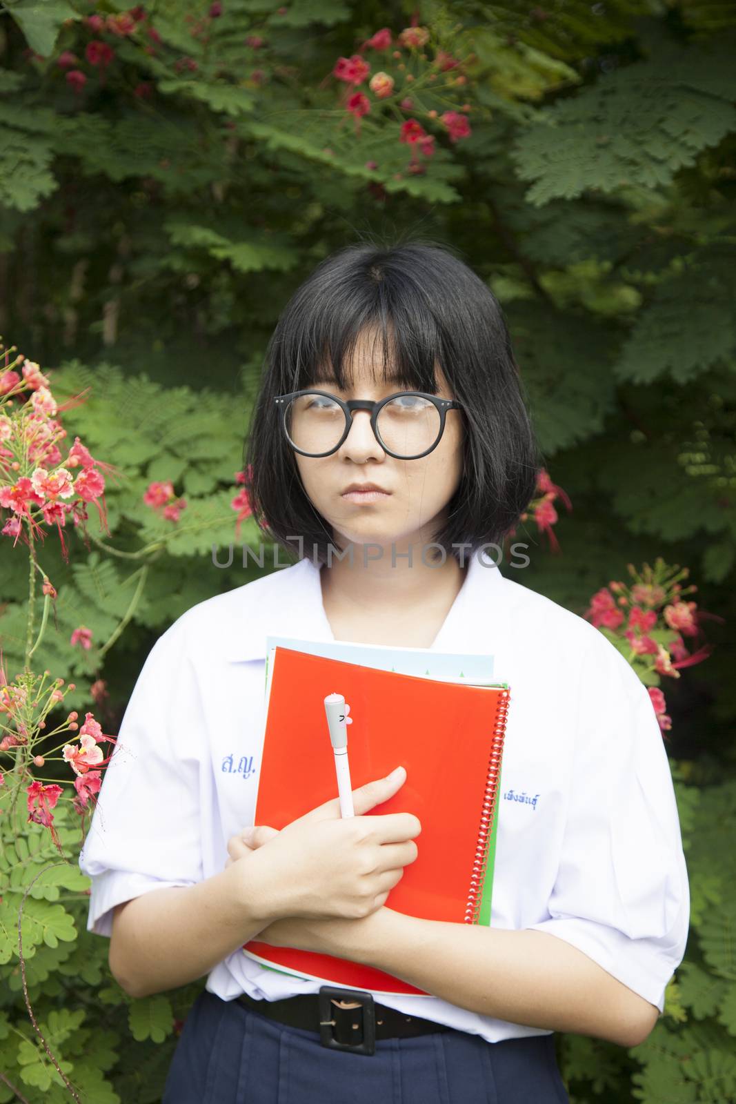 Schoolgirl with wear glasses looks sad. Standing near a tree with red flowers. Within the park