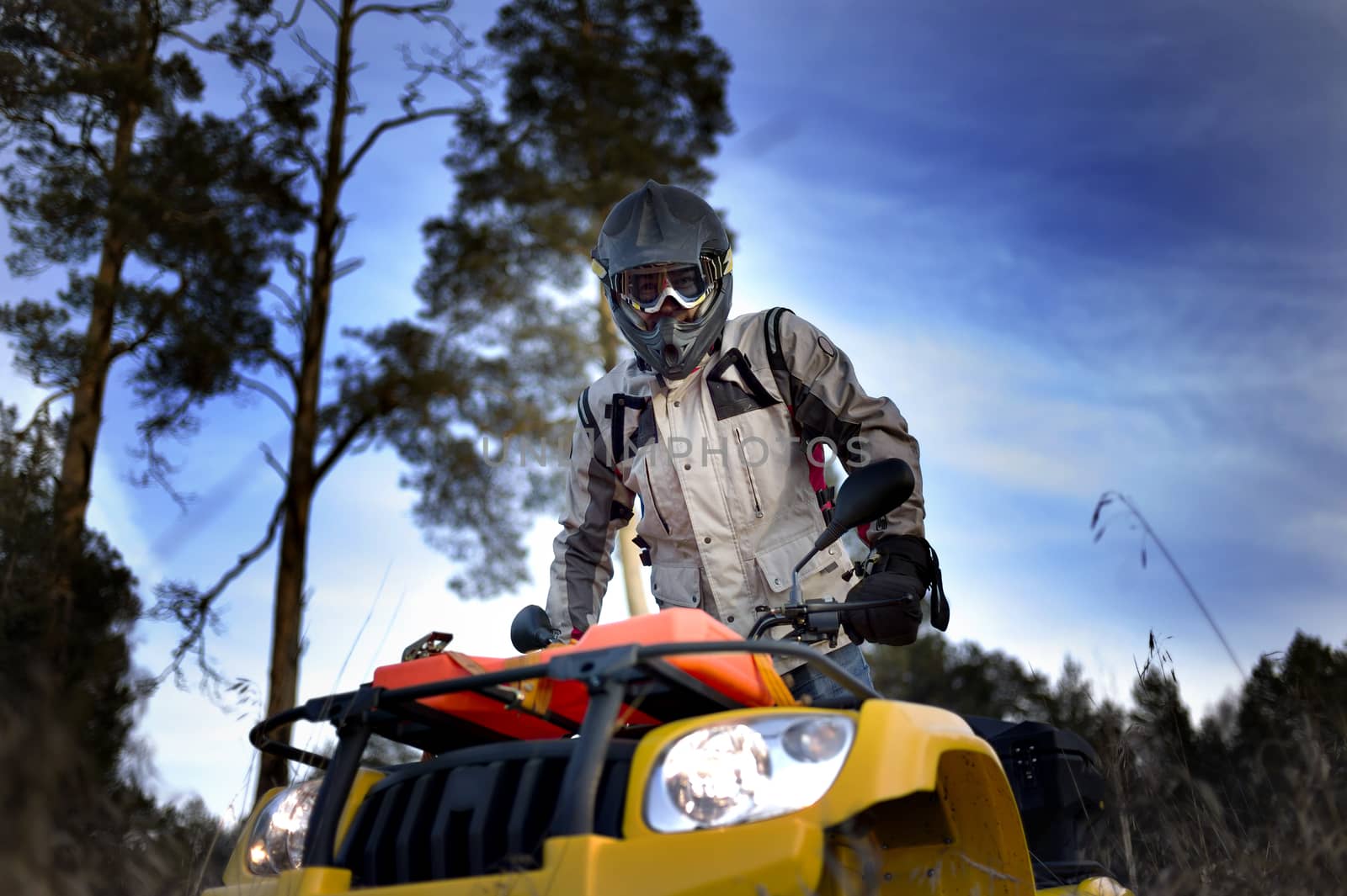 Horizontal close-up of a man in helmet and safety goggles looking into the camera while sitting on quad bike against vivid blue sky.