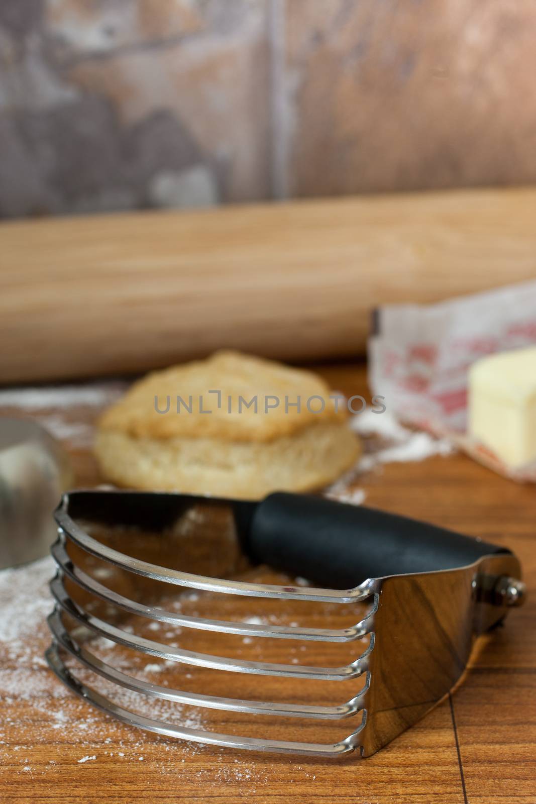 A chrome pastry cutter sits on a counter top with rolling pin, butter, flour, and a biscuit on the background.