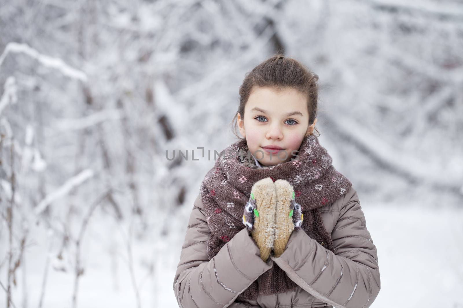 Close-up portrait of a little girl in brown jacket and knit scarf and hat on a background of a snow park