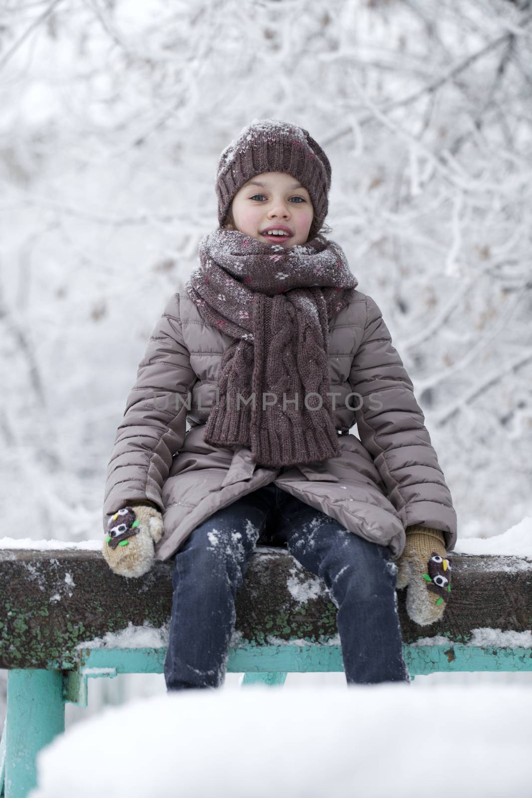 Close-up portrait of a little girl in brown jacket and knit scarf and hat on a background of a snow park