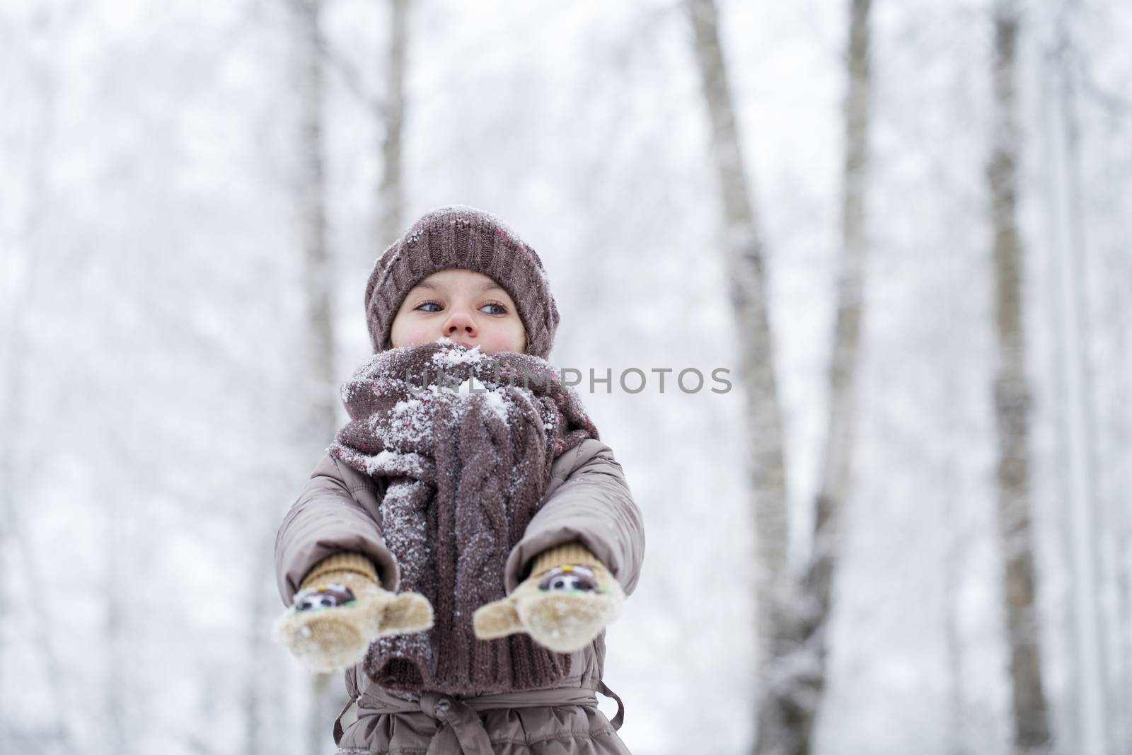 Close-up portrait of a little girl in brown jacket and knit scarf and hat on a background of a snow park