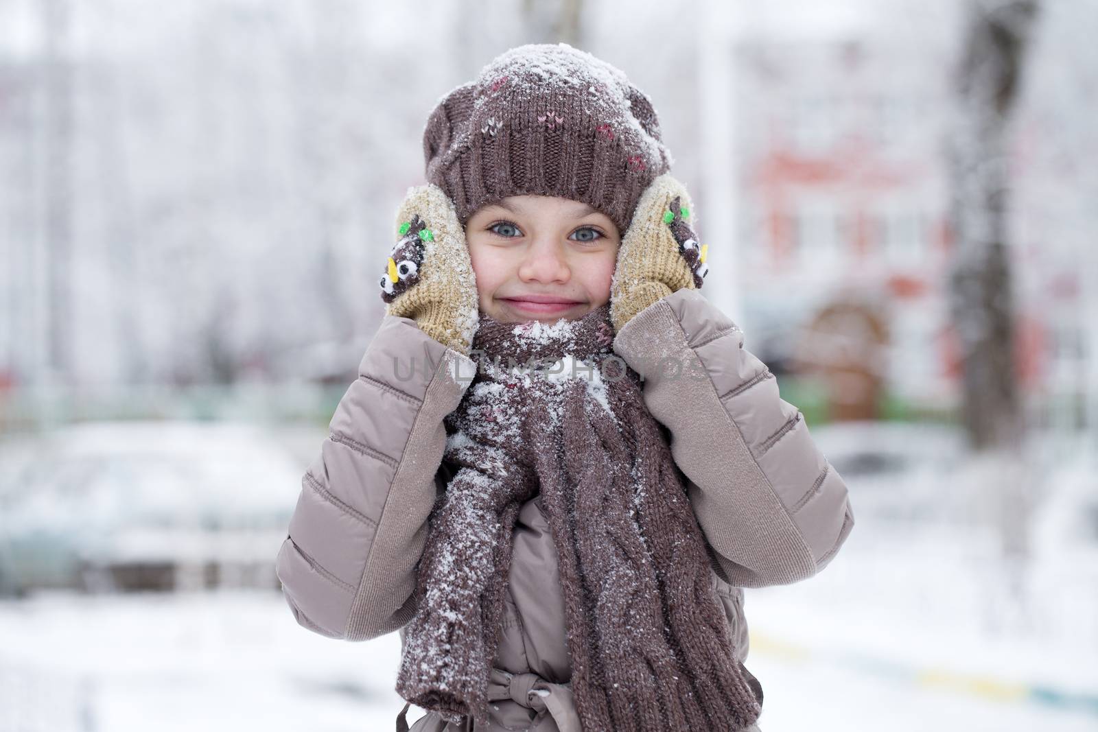 Close-up portrait of a little girl in brown jacket and knit scarf and hat on a background of a snow park