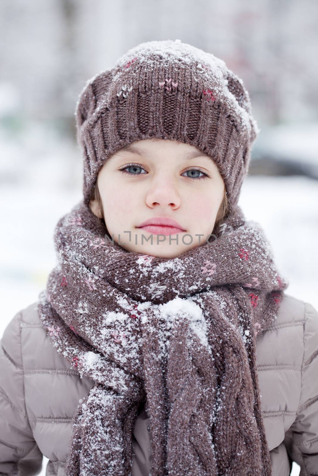 Close-up portrait of a little girl in brown jacket and knit scarf and hat on a background of a snow park