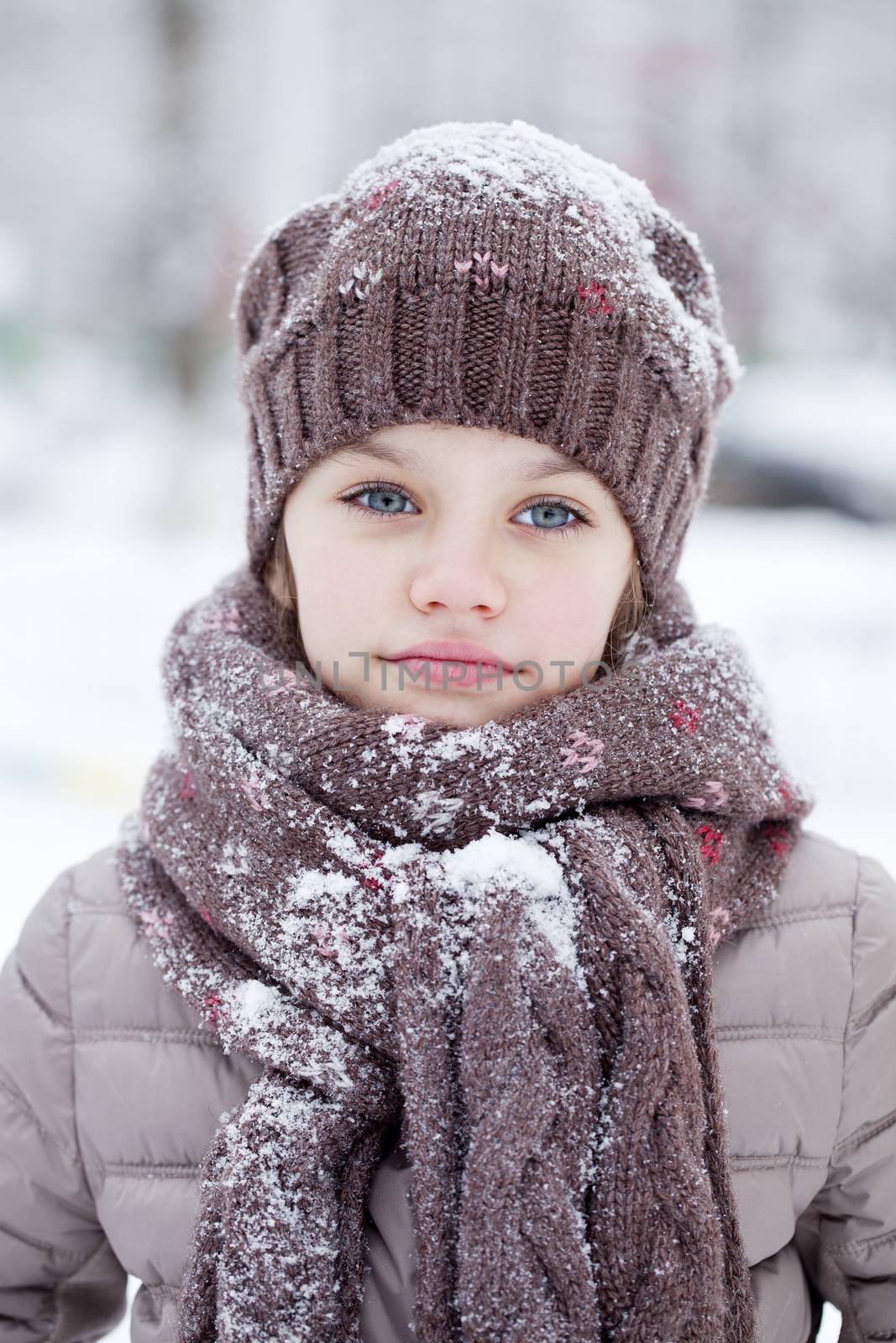 Close-up portrait of a little girl in brown jacket and knit scarf and hat on a background of a snow park