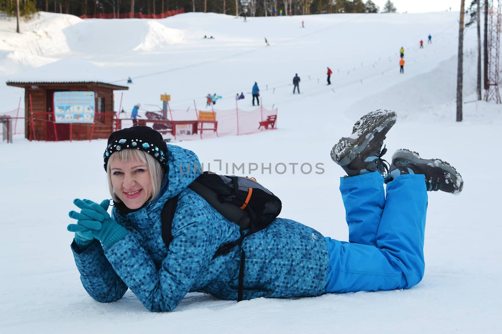 The woman in winter clothes lies on snow against the ski slope and smiles