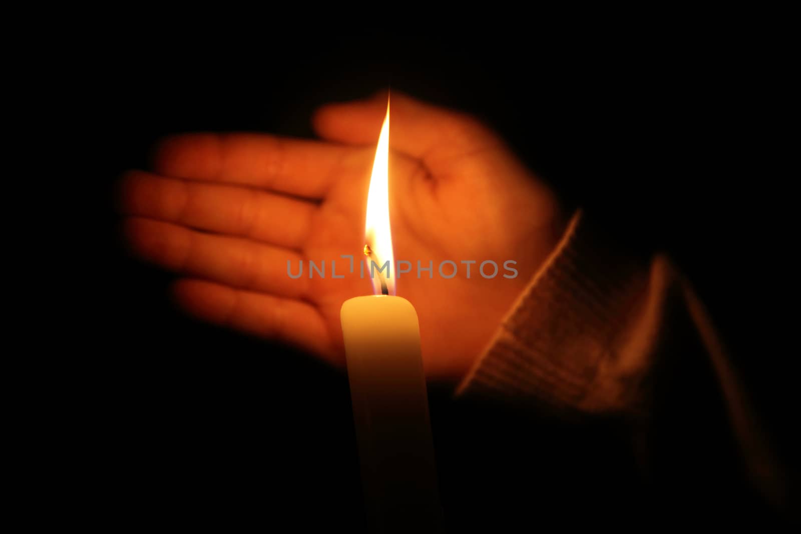 hand protects the flame from a candle burning on a dark background