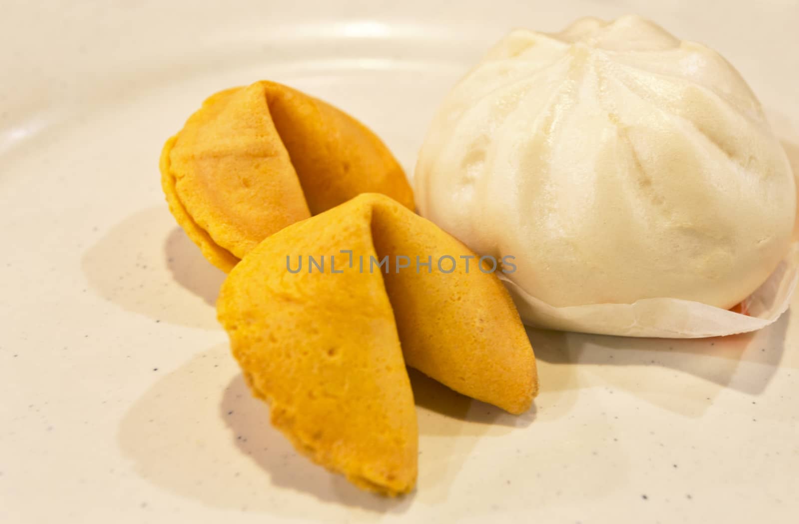 One Chinese steamed bun next to two fortune cookies on a rustic,  beige stoneware plate.  