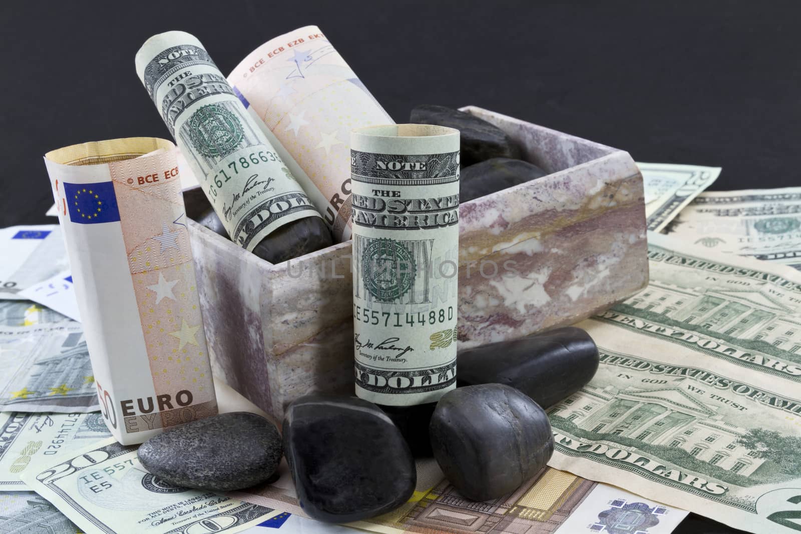 European euros and American dollars sit on a bed of currency within a marble box accented with black rocks suggesting difficult financial trends in international banking, investment, and business.  Black background and upper area copy space. 