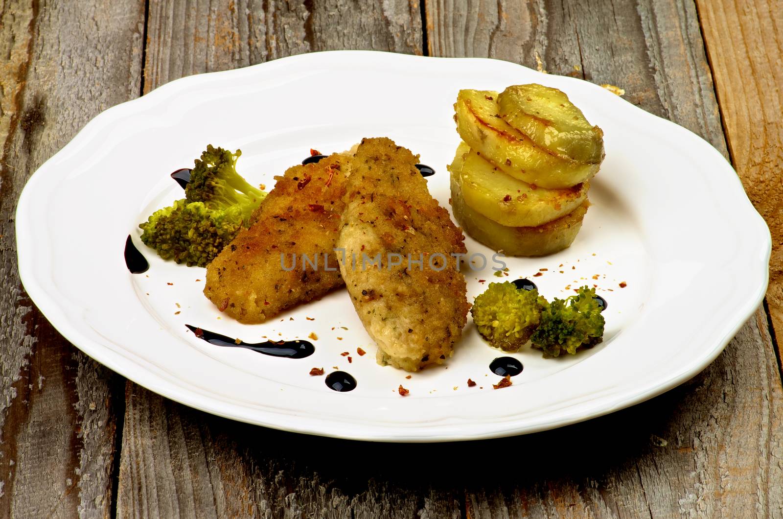 Delicious Fried Chicken Strips in Herb Breading Served with Steamed Broccoli, Roasted Potato Slices and Balsamic Sauce on White Plate isolated on Rustic Wooden background