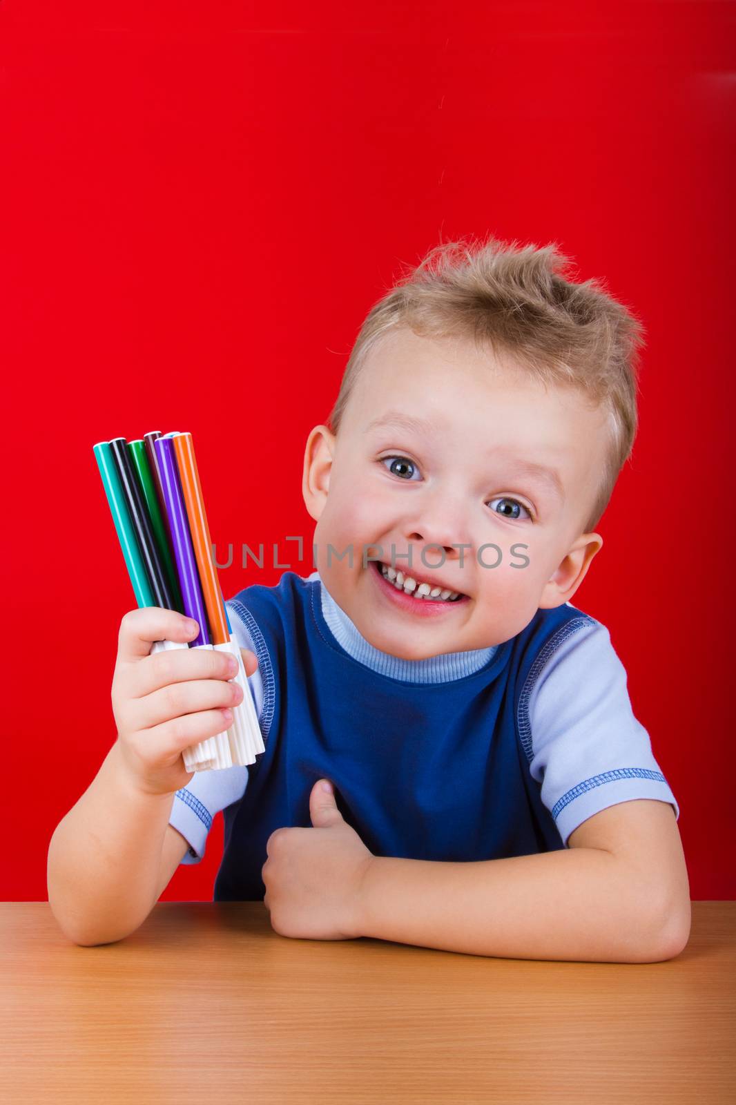 Small boy is sitting at the table and holding a colorful markers. On a red background