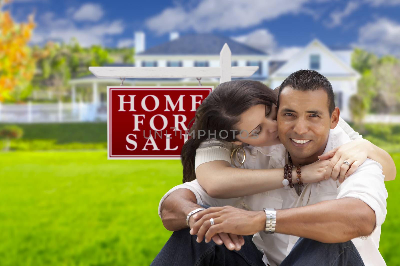 Young Happy Hispanic Young Couple in Front of Their New Home and For Sale Real Estate Sign.