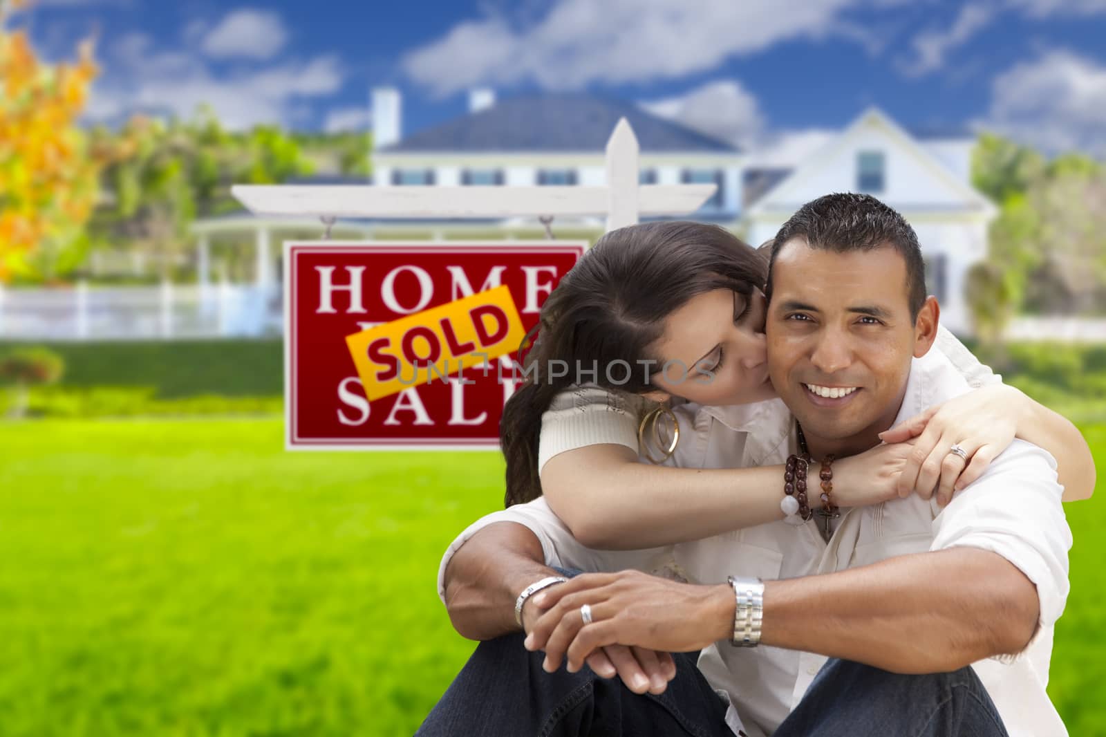 Young Happy Hispanic Young Couple in Front of Their New Home and Sold For Sale Real Estate Sign.