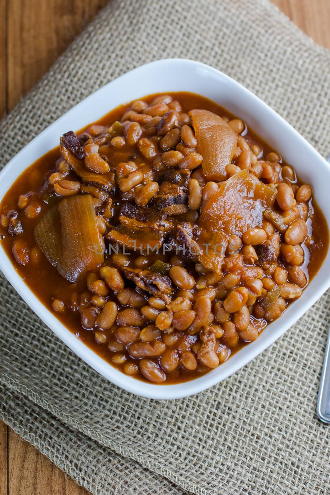 Rustic baked beans in a white bowl on a burlap placemat and wooden table.