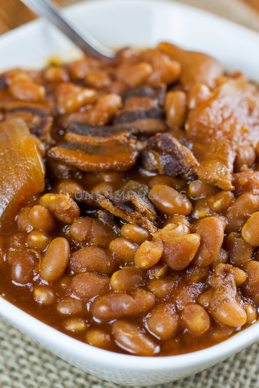 Rustic baked beans in a white bowl on a burlap placemat and wooden table.