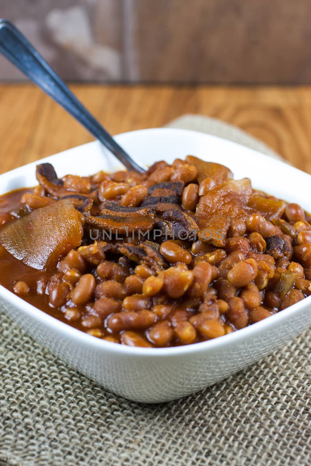 Rustic baked beans in a white bowl on a burlap placemat and wooden table.