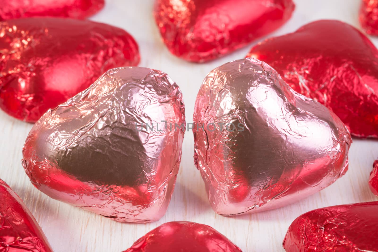 Two chocolate hearts, in pink wrapping foil, close together and surrounded by many other chocolate hearts in red wrapping foil.