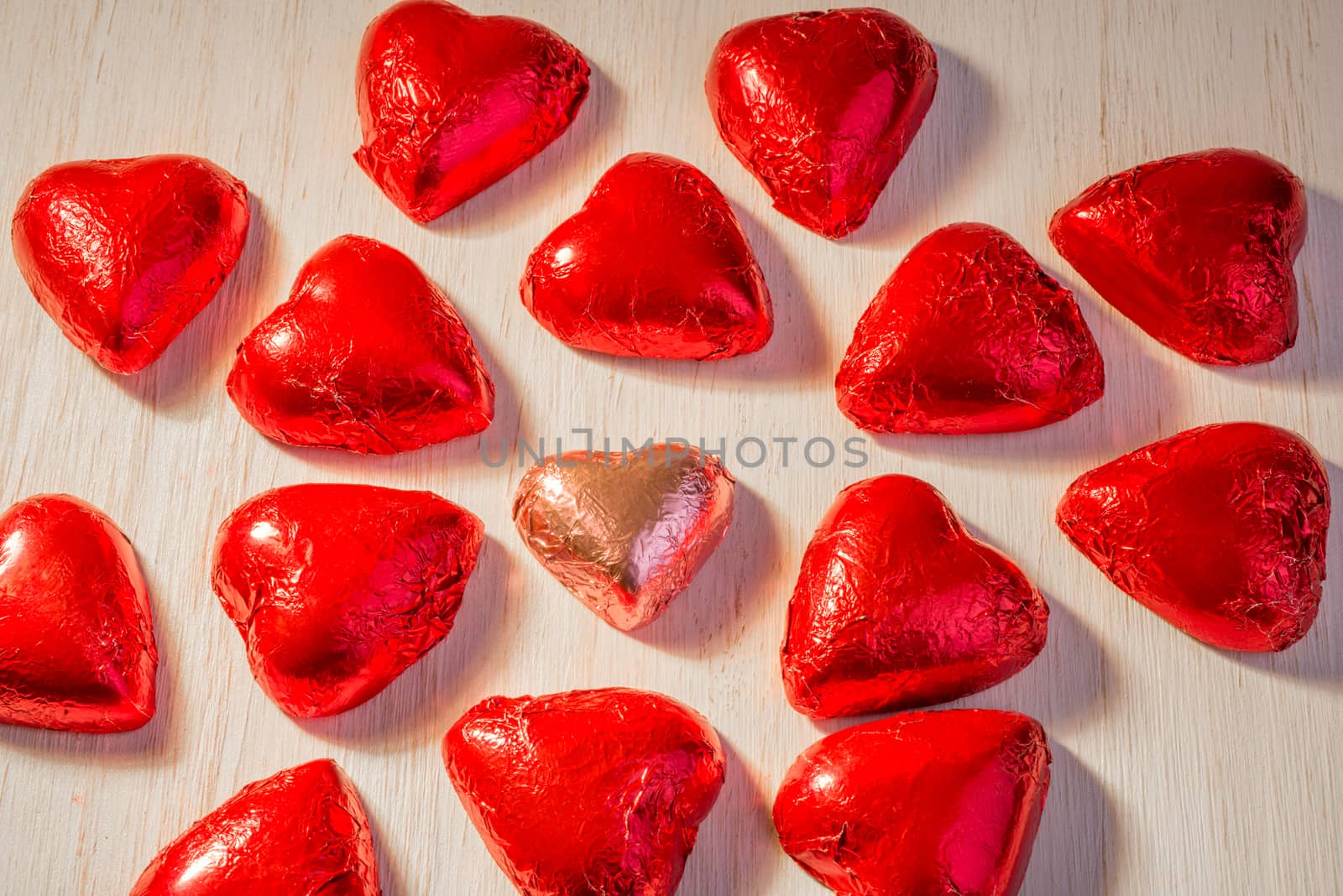 One chocoalte heart in pink wrapping paper, surrounded by many hearts in red wrapping paper.