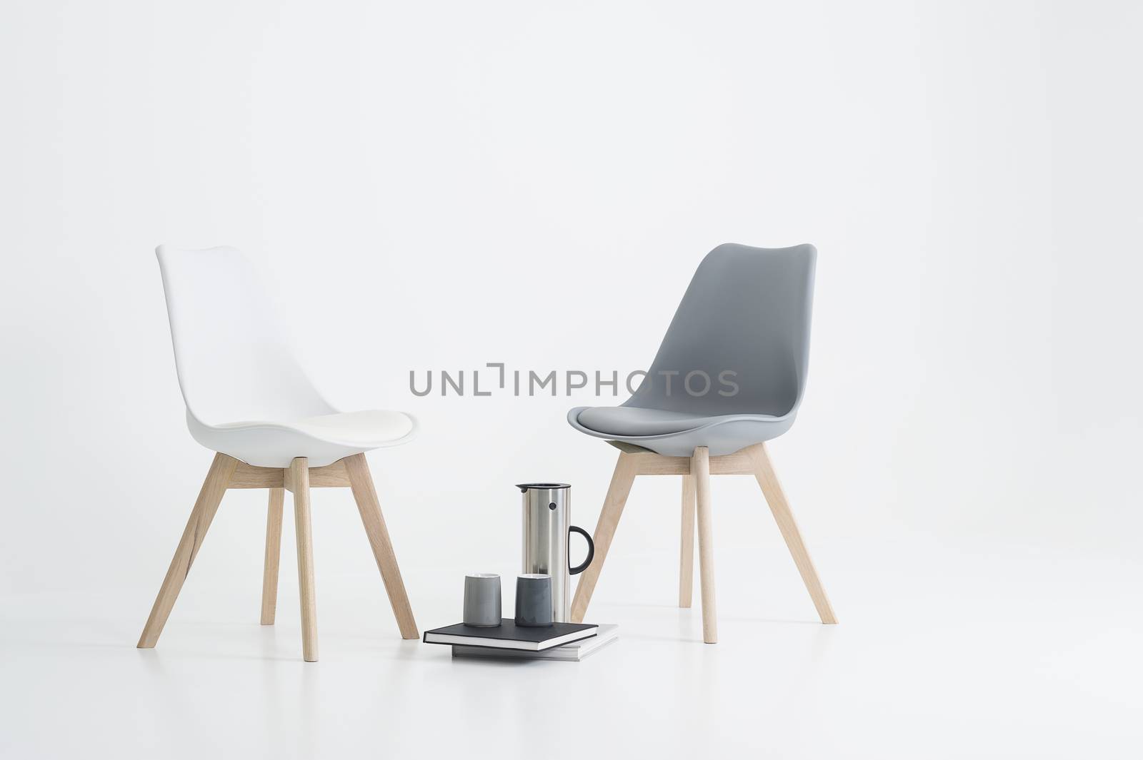 Two modern chairs with a serving of coffee in a stylish flask with two mugs resting on hardcover books on the floor in between, over white