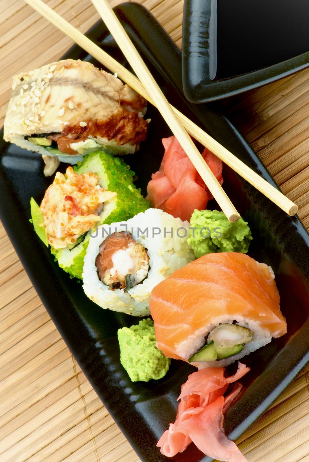 Set of Various Maki Sushi with Salmon, Eel, Green Caviar and Crab on Black Plate with Soy Sauce and Pair of Chopsticks on Straw mat background
