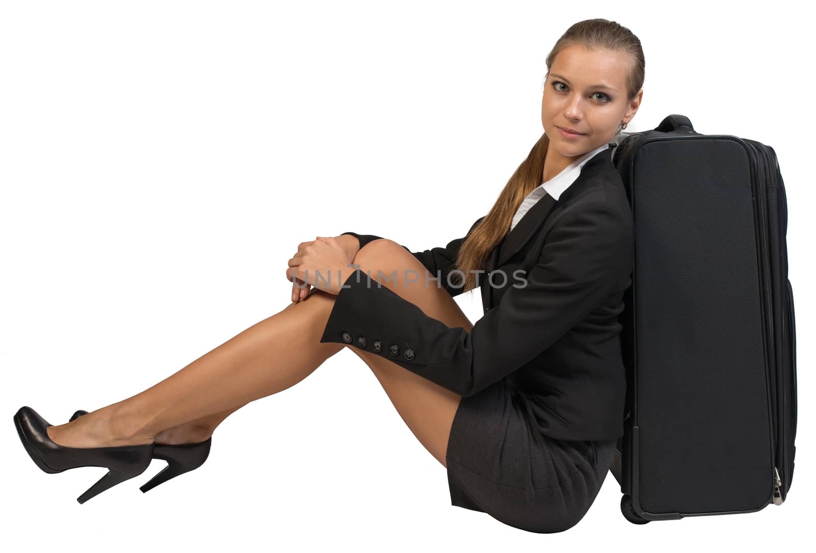 Businesswoman sitting next to side view suitcase, hugging her knees, looking at camera. Isolated over white background