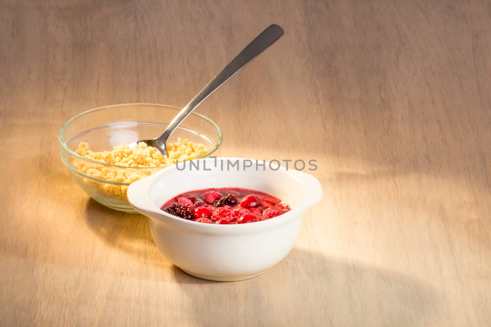 Berry mix in ceramic bowl with crumble in a glass bowl together with a spoon on table.