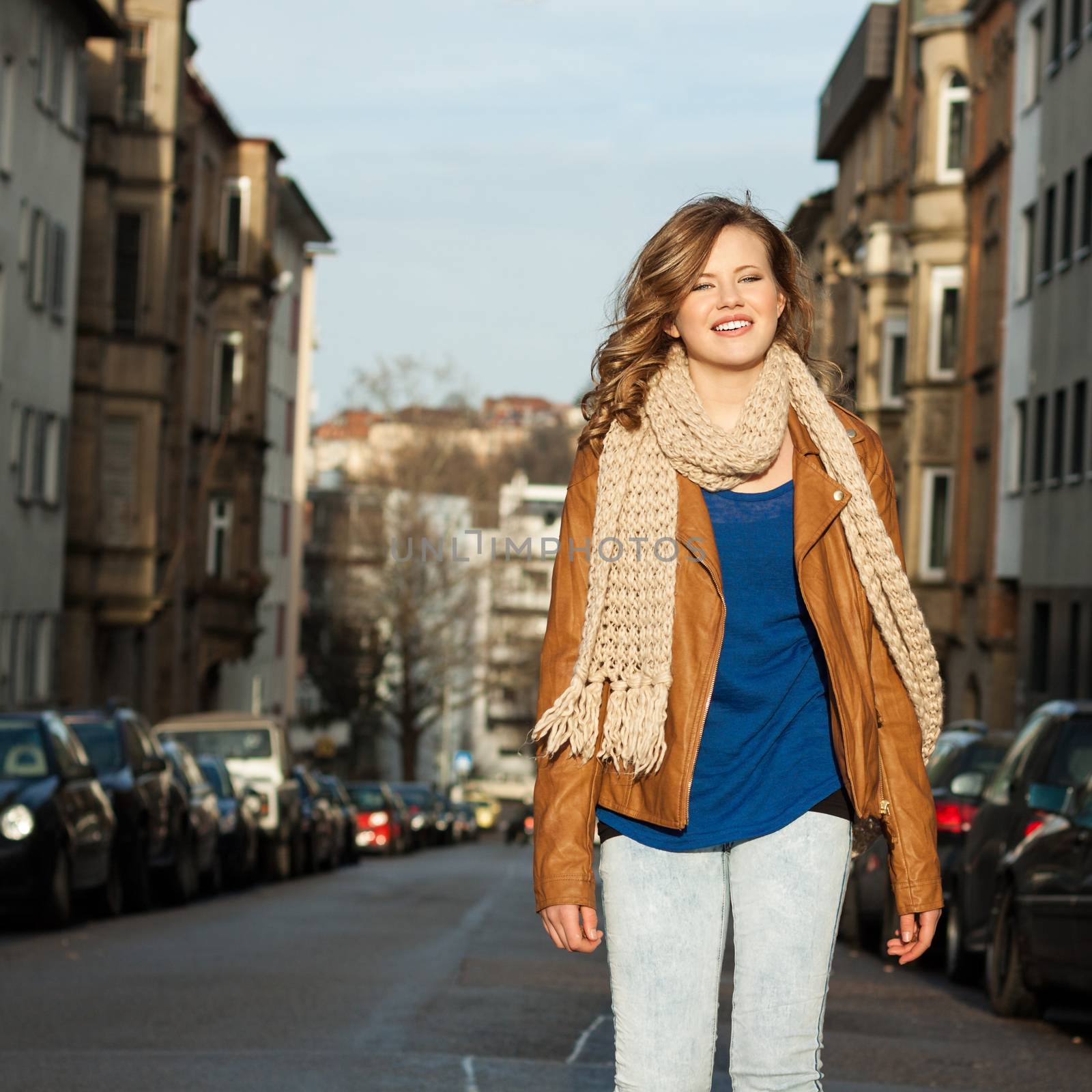 Beautiful trendy young woman with a lovely smile in a long flowing scarf standing in a shaft of sunlight in an urban street with parked cars and townhouses