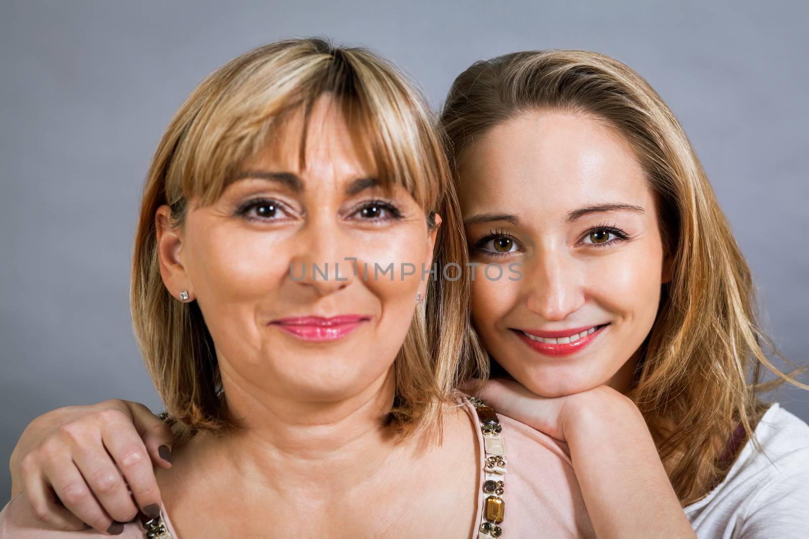 Playful beautiful young mother and her teenage daughter posing together with the young girl peeking out to the side with a happy grin, isolated on a grey studio background