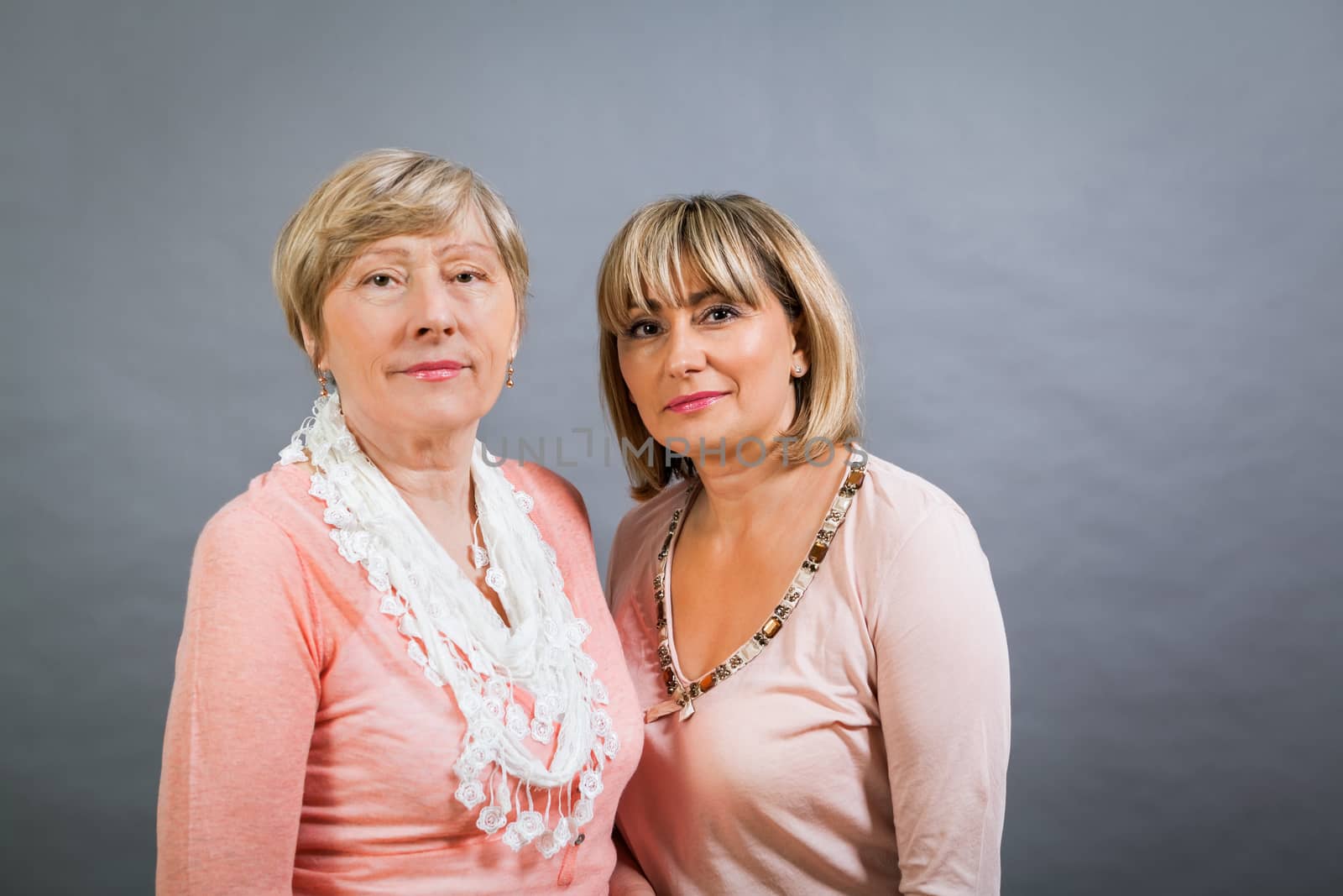 Attractive stylish blond senior lady with her beautiful middle-aged daughter posing together with her hands on her shoulders smiling at the camera on a grey studio background