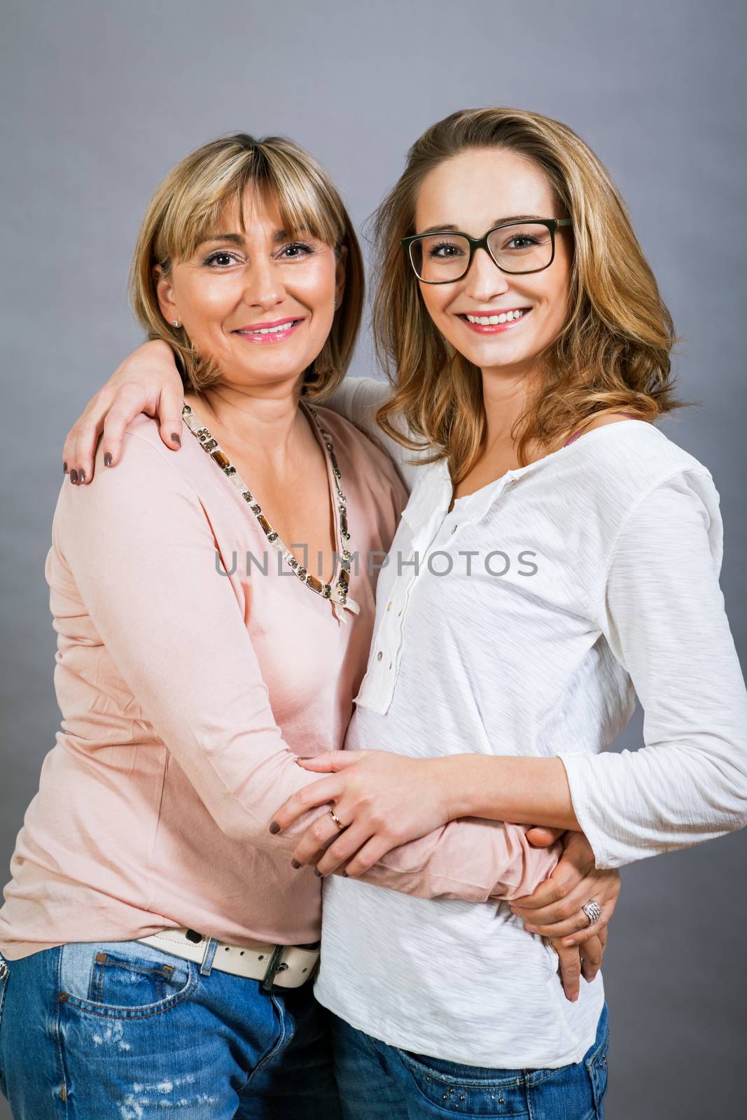 Playful beautiful young mother and her teenage daughter posing together with the young girl peeking out to the side with a happy grin, isolated on a grey studio background