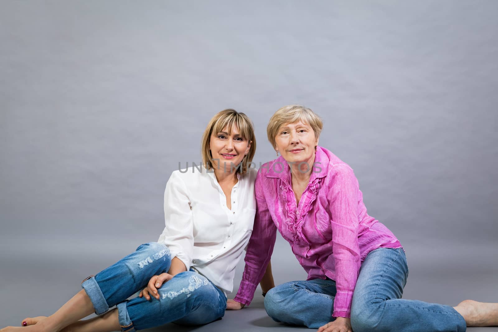 Attractive stylish blond senior lady with her beautiful middle-aged daughter posing together with her hands on her shoulders smiling at the camera on a grey studio background