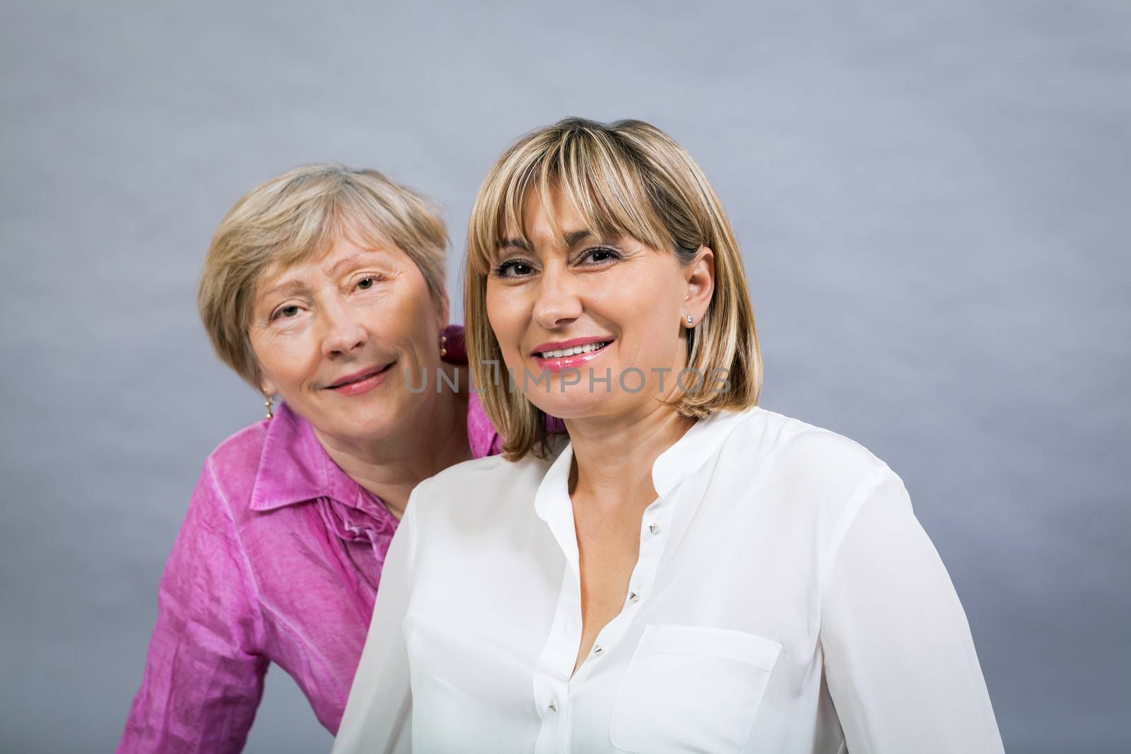 Attractive stylish blond senior lady with her beautiful middle-aged daughter posing together with her hands on her shoulders smiling at the camera on a grey studio background