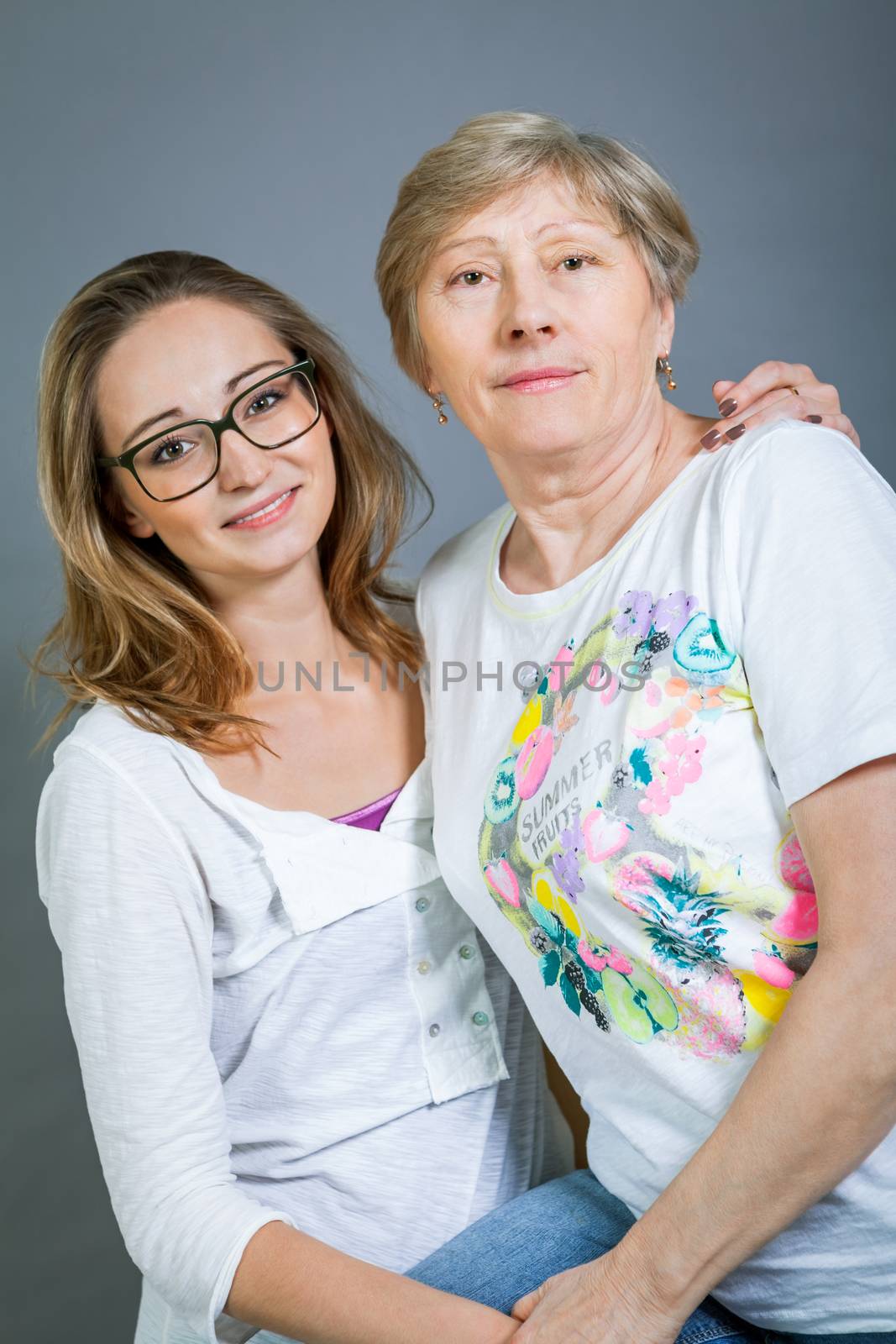 Loving grandmother and teenage granddaughter posing for a portrait holding hands and smiling at the camera, on a grey studio background