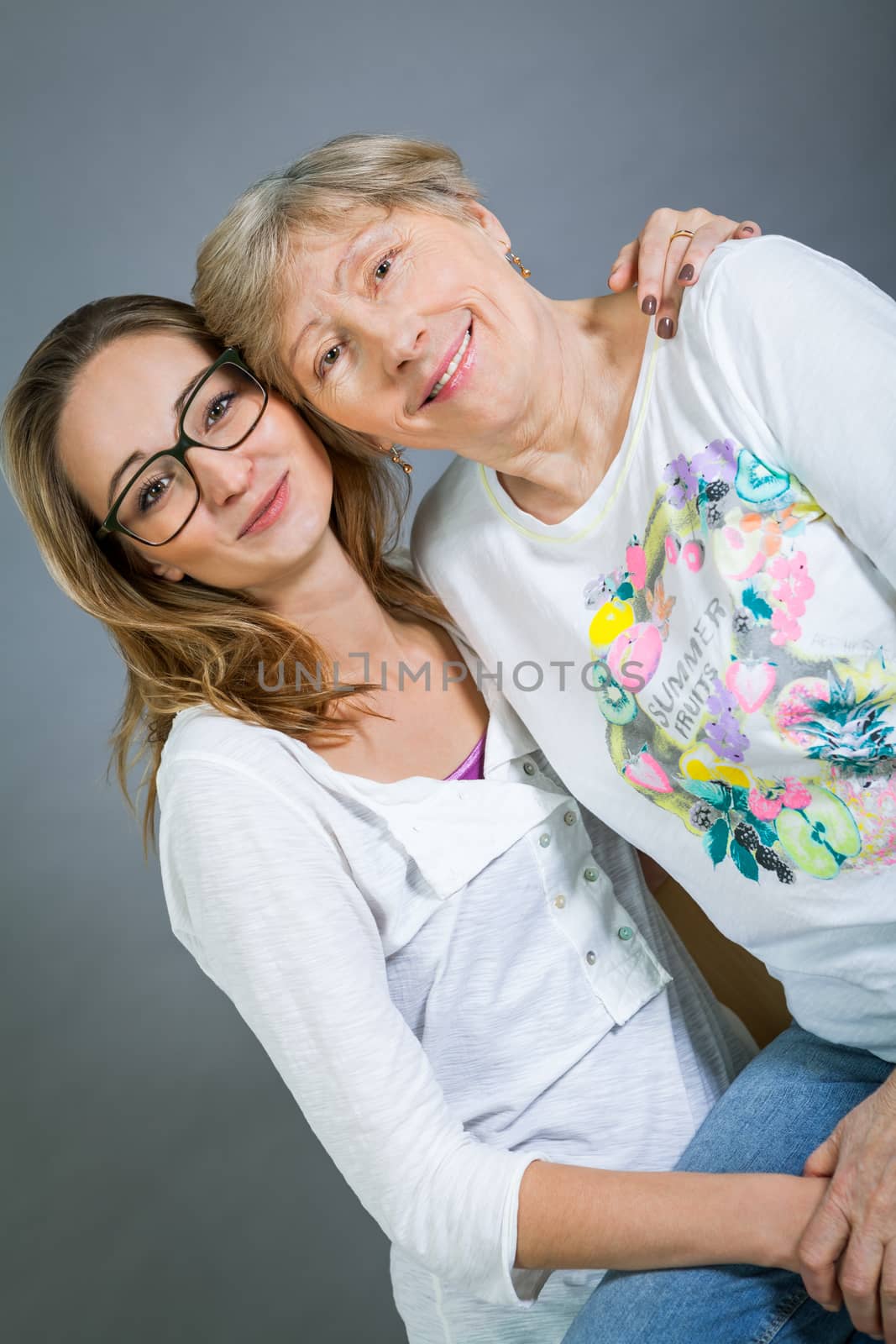 Loving grandmother and teenage granddaughter posing for a portrait holding hands and smiling at the camera, on a grey studio background