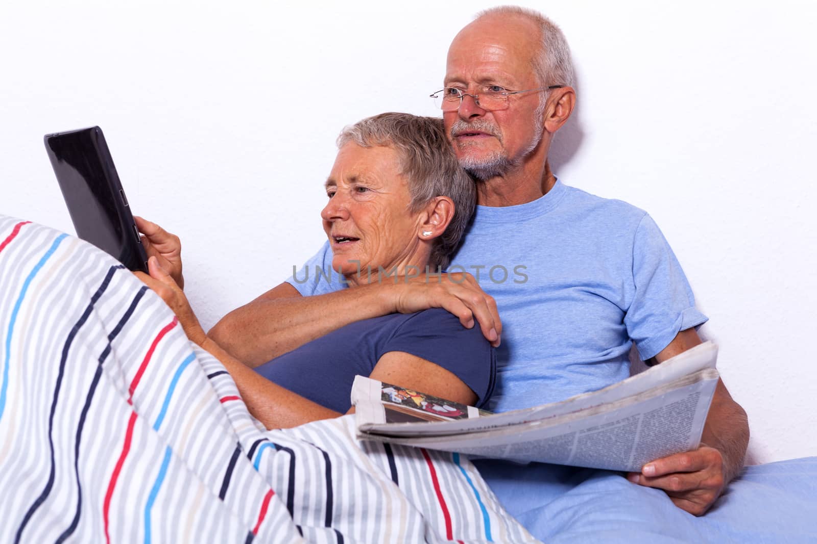 Senior Couple Relaxing in Bed with Tablet Computer and Newspaper