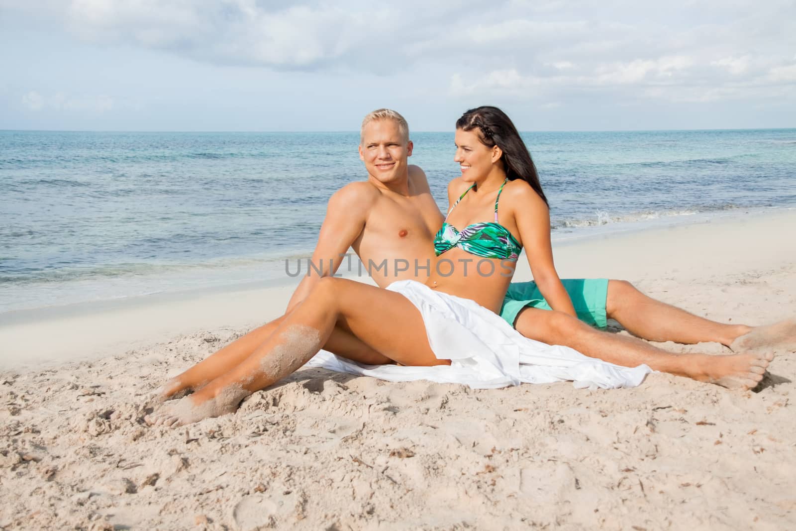 Happy attractive affectionate young couple sunbathing together on the beach in their swimwear while enjoying a tropical summer holiday during the annual vacation