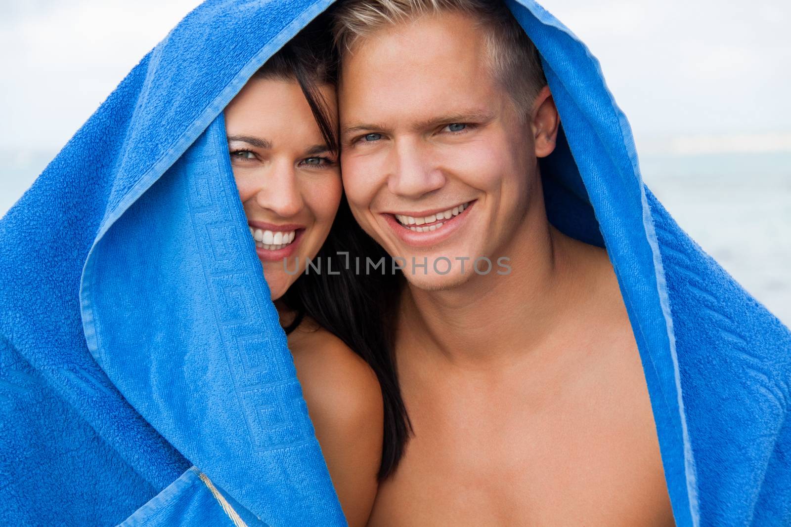 Cheerful Caucasian young happy couple made of a blond handsome man and an attractive brunette woman, with a blue towel covering their heads