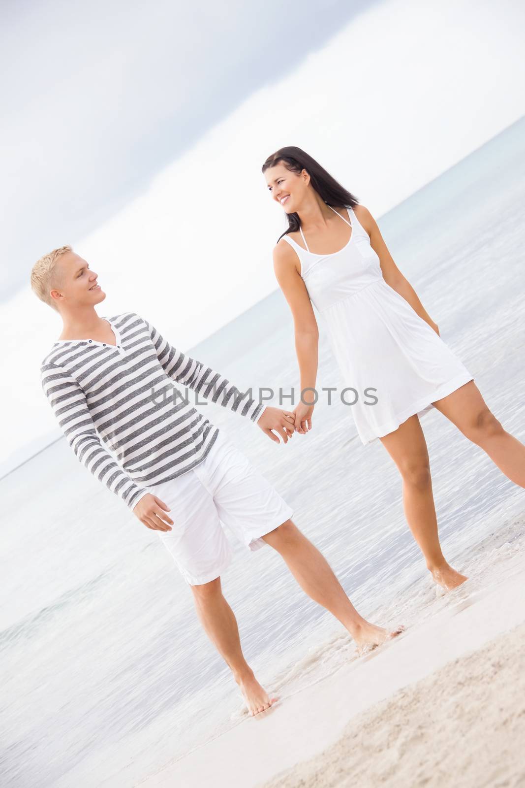 Caucasian happy young couple holding hands while walking barefoot on the beach in a romantic travel destination