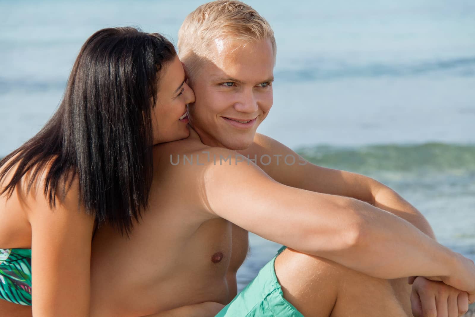 Happy attractive affectionate young couple sunbathing together on the beach in their swimwear while enjoying a tropical summer holiday during the annual vacation