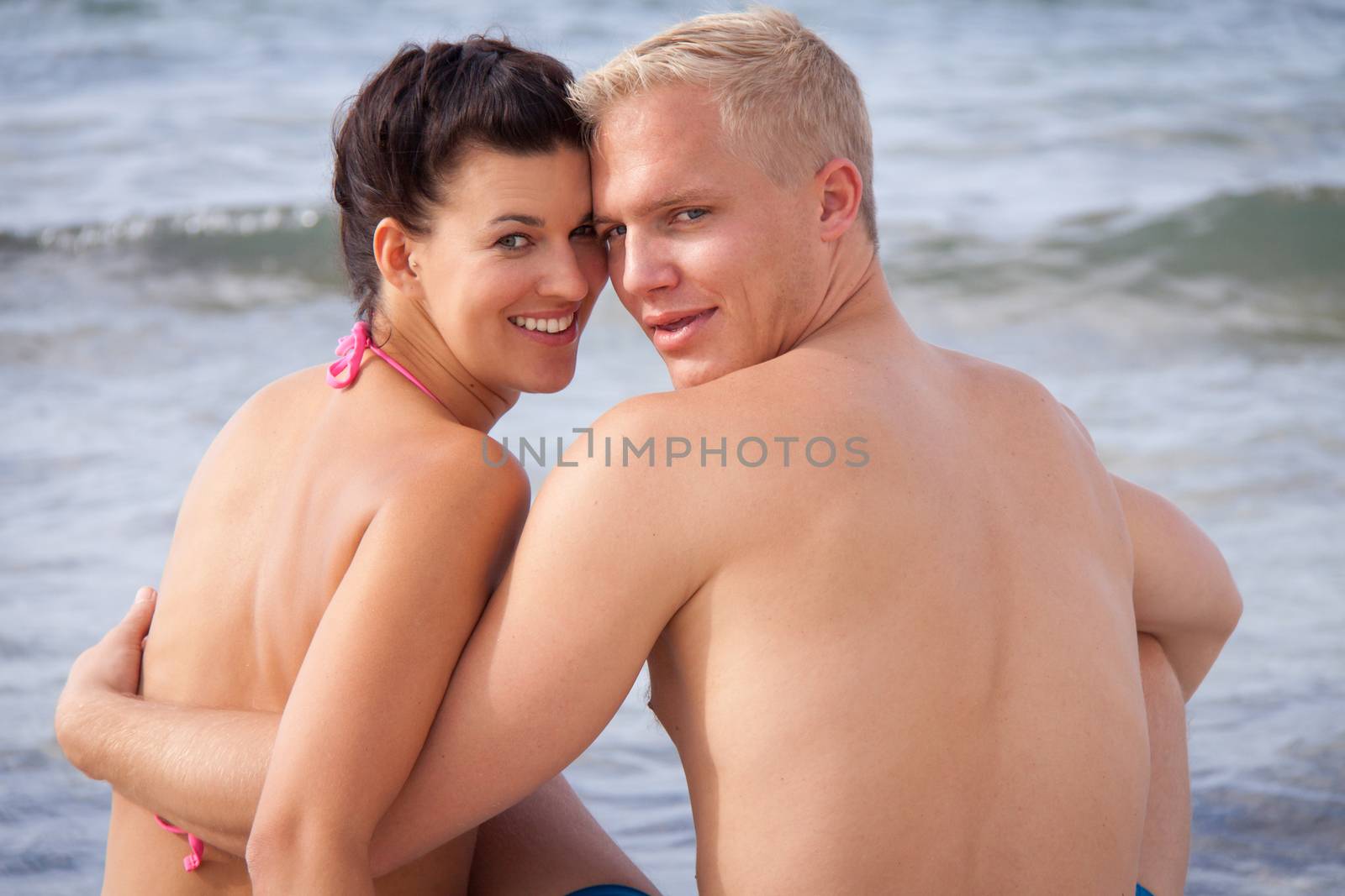 Happy romantic young couple enjoying a date at the seaside sitting arm in arm overlooking the ocean and looking back at the camera with charming happy smiles