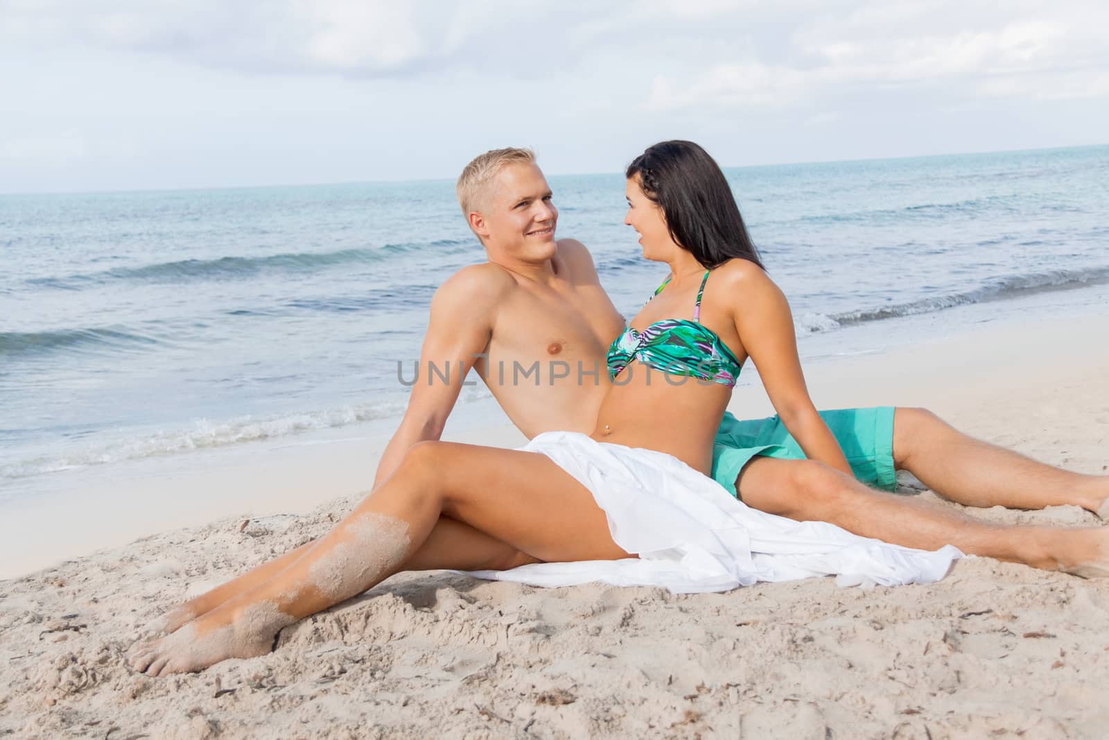 Happy attractive affectionate young couple sunbathing together on the beach in their swimwear while enjoying a tropical summer holiday during the annual vacation